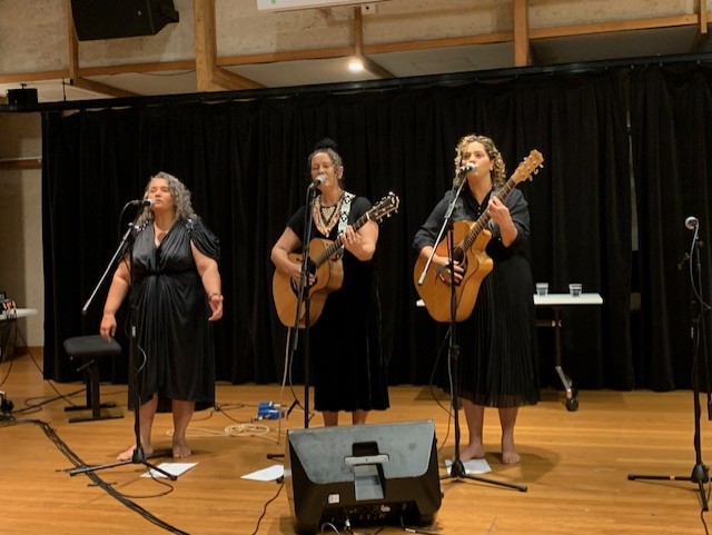 Three people singing into microphones. Two of the singers are also playing guitars.