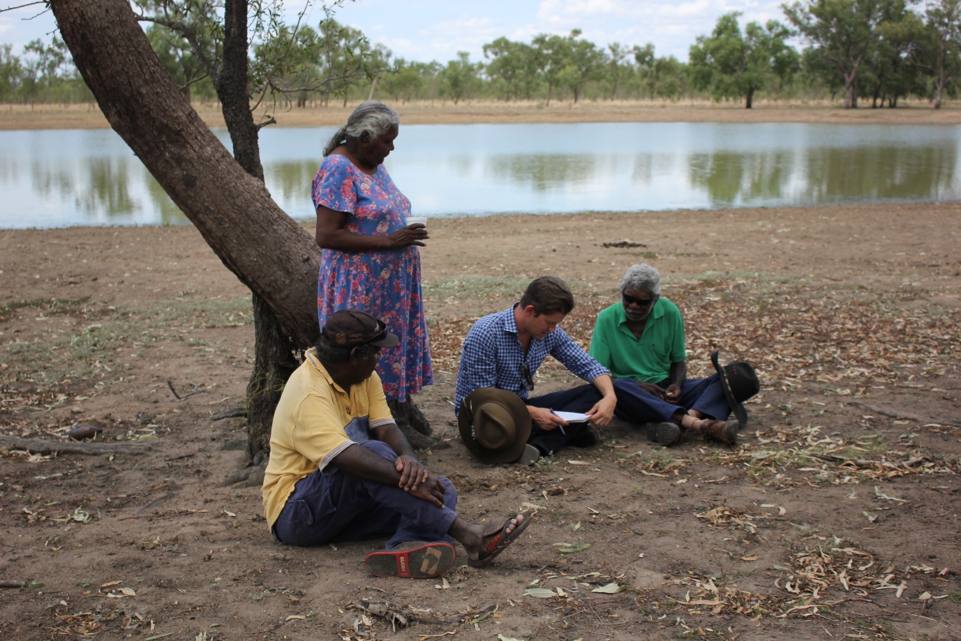 Three people seated on the ground and one person next to a tree in front of the water. The author is holding a notebook and pen.