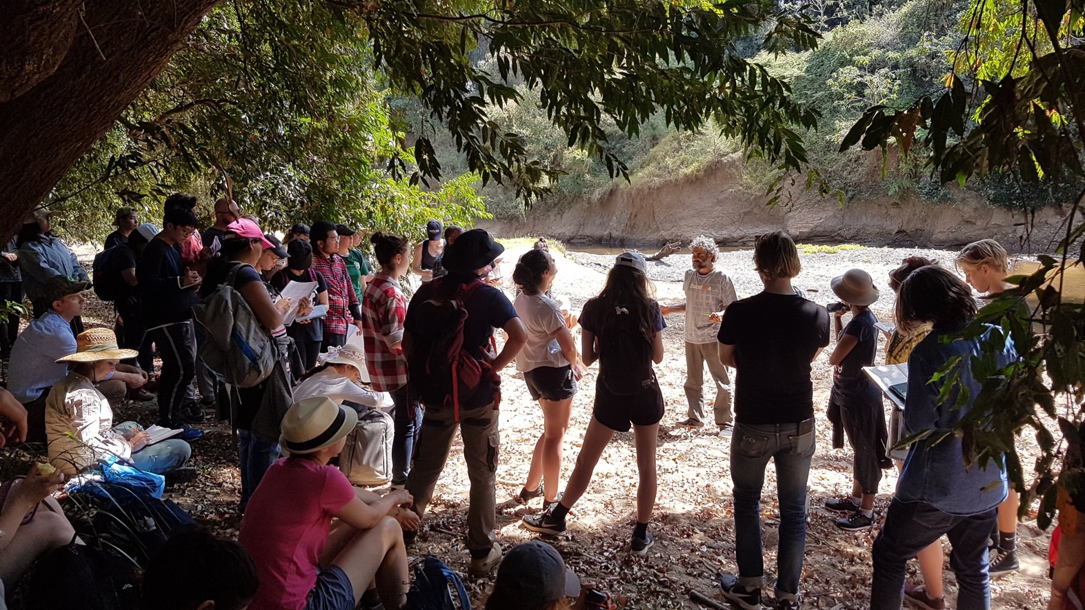 A person standing on a river bank under trees, talking to a group of people.