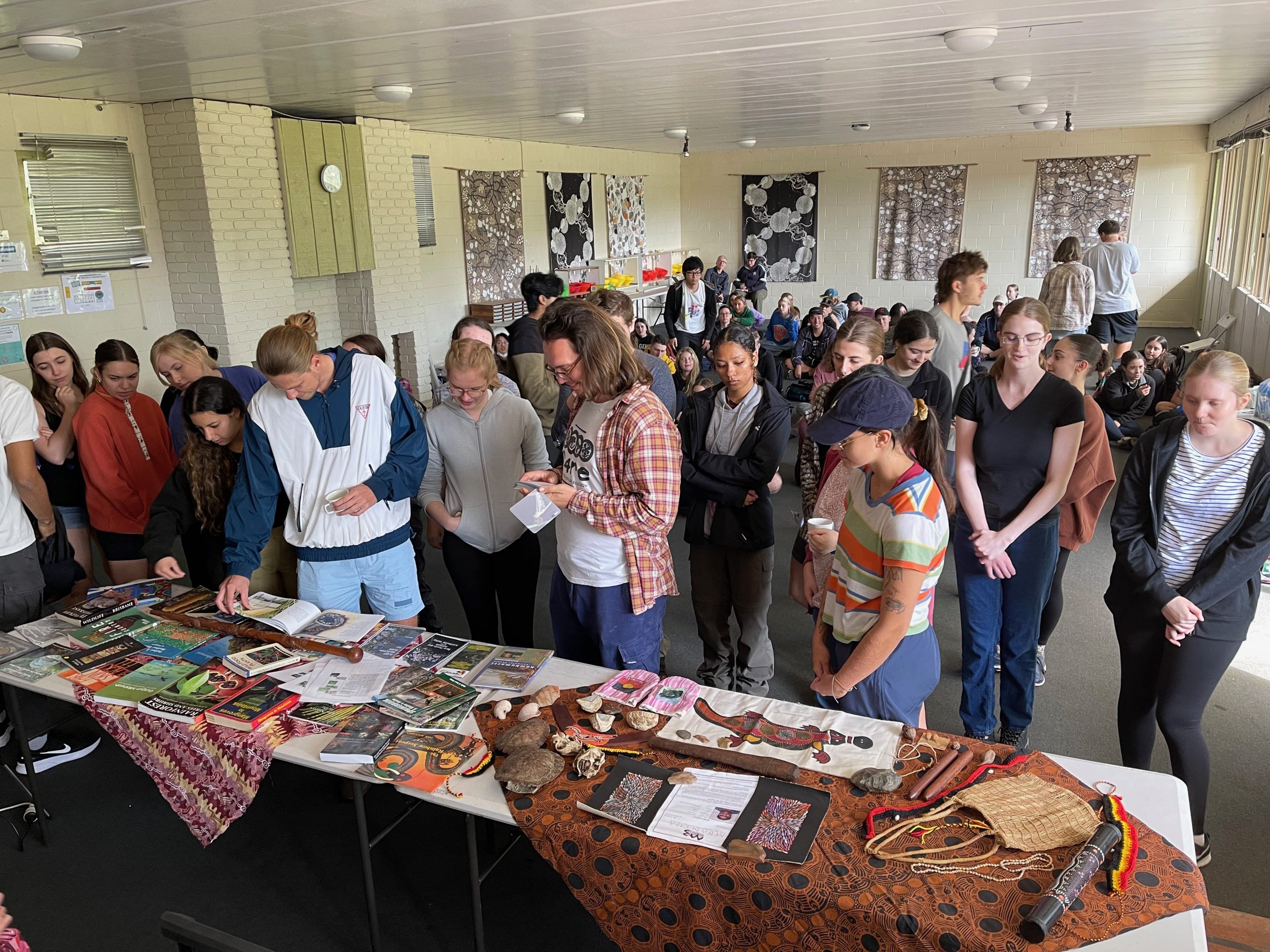Students standing in a room looking at a table with display items. Other students sit on the floor behind them.