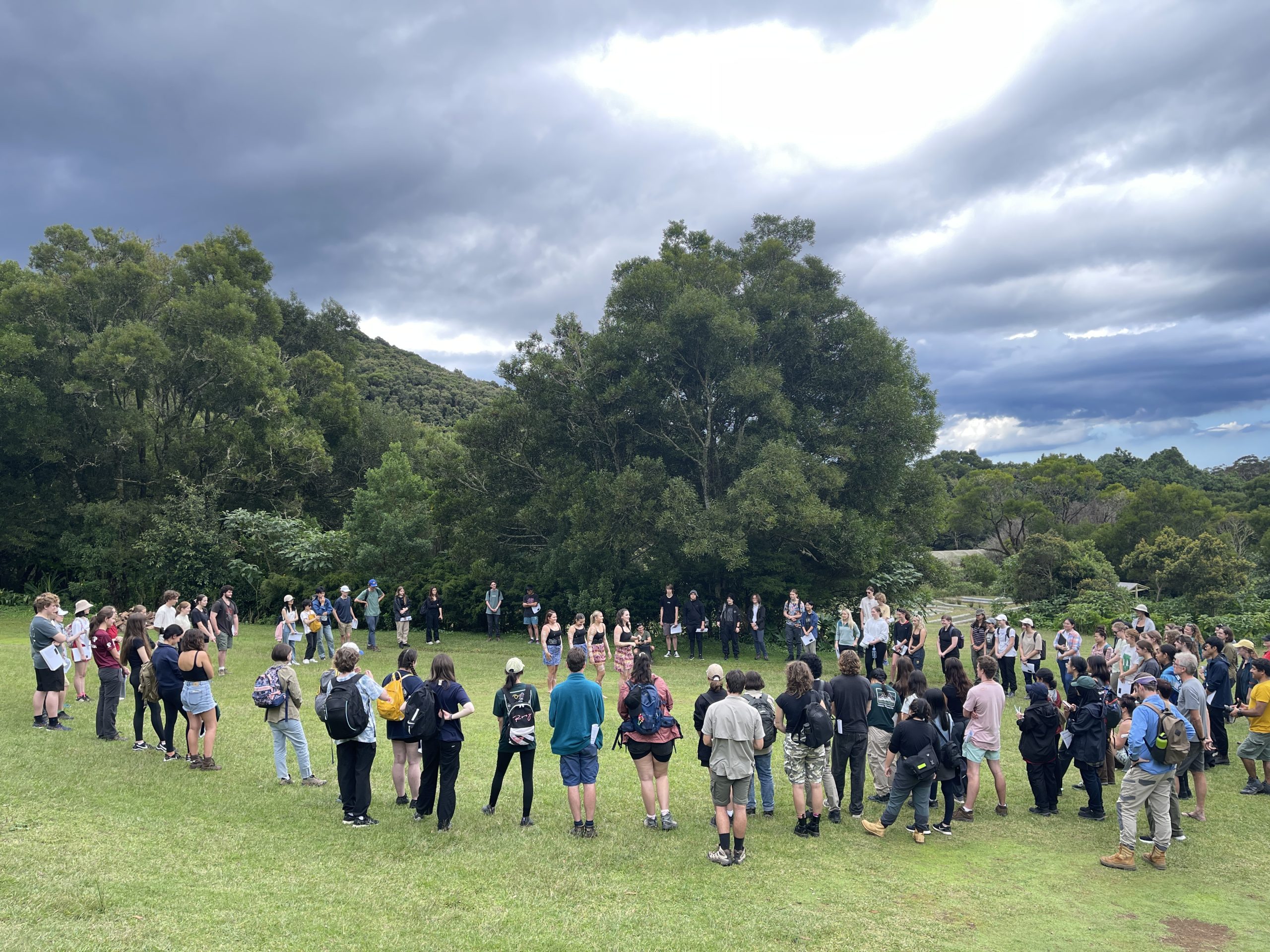 The students stand in a circle on the grass with trees behind. The performers are standing in the centre of the circle.