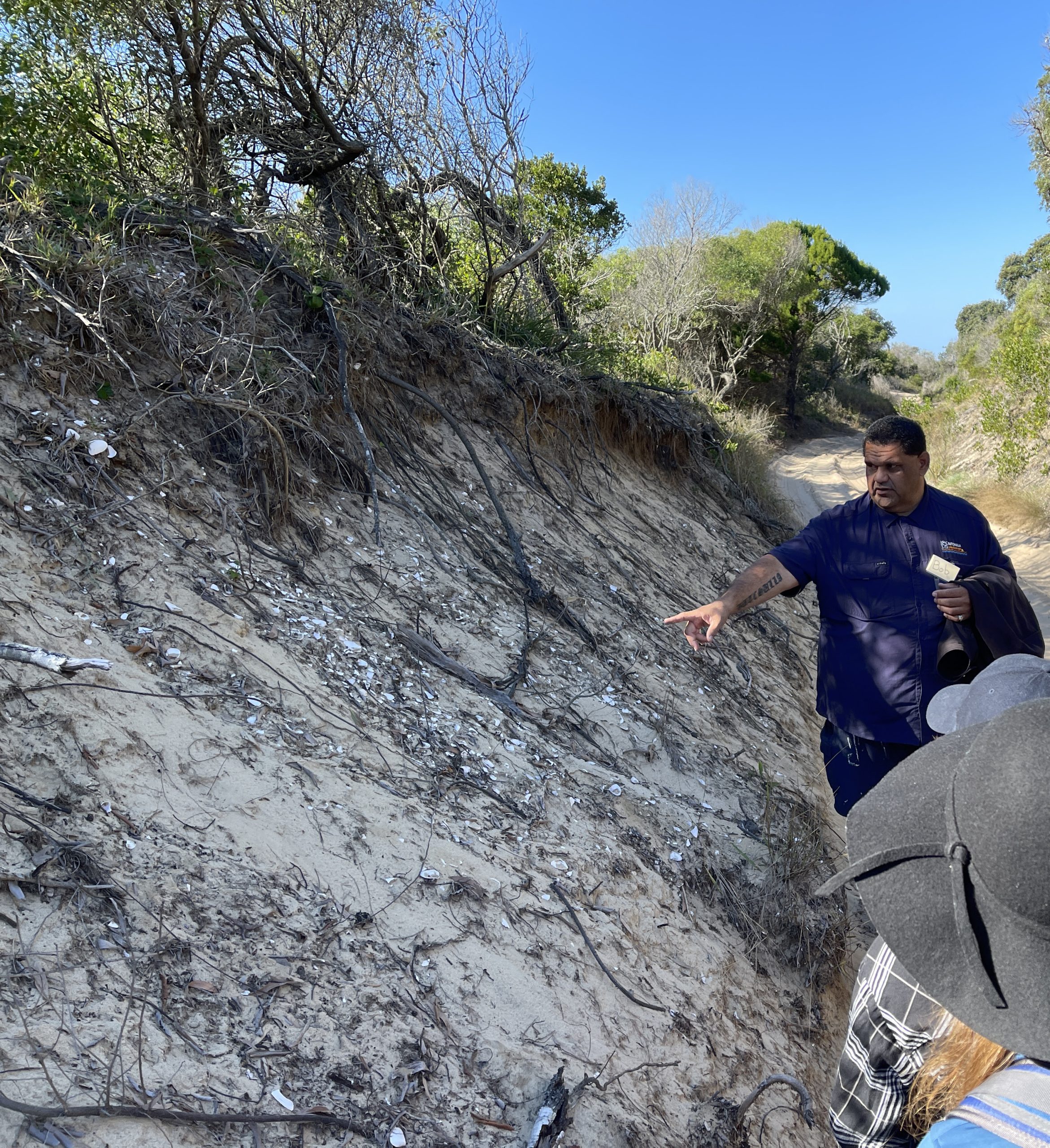 Butchulla Land and Sea Ranger Bob Broome points to the midden with people standing nearby.