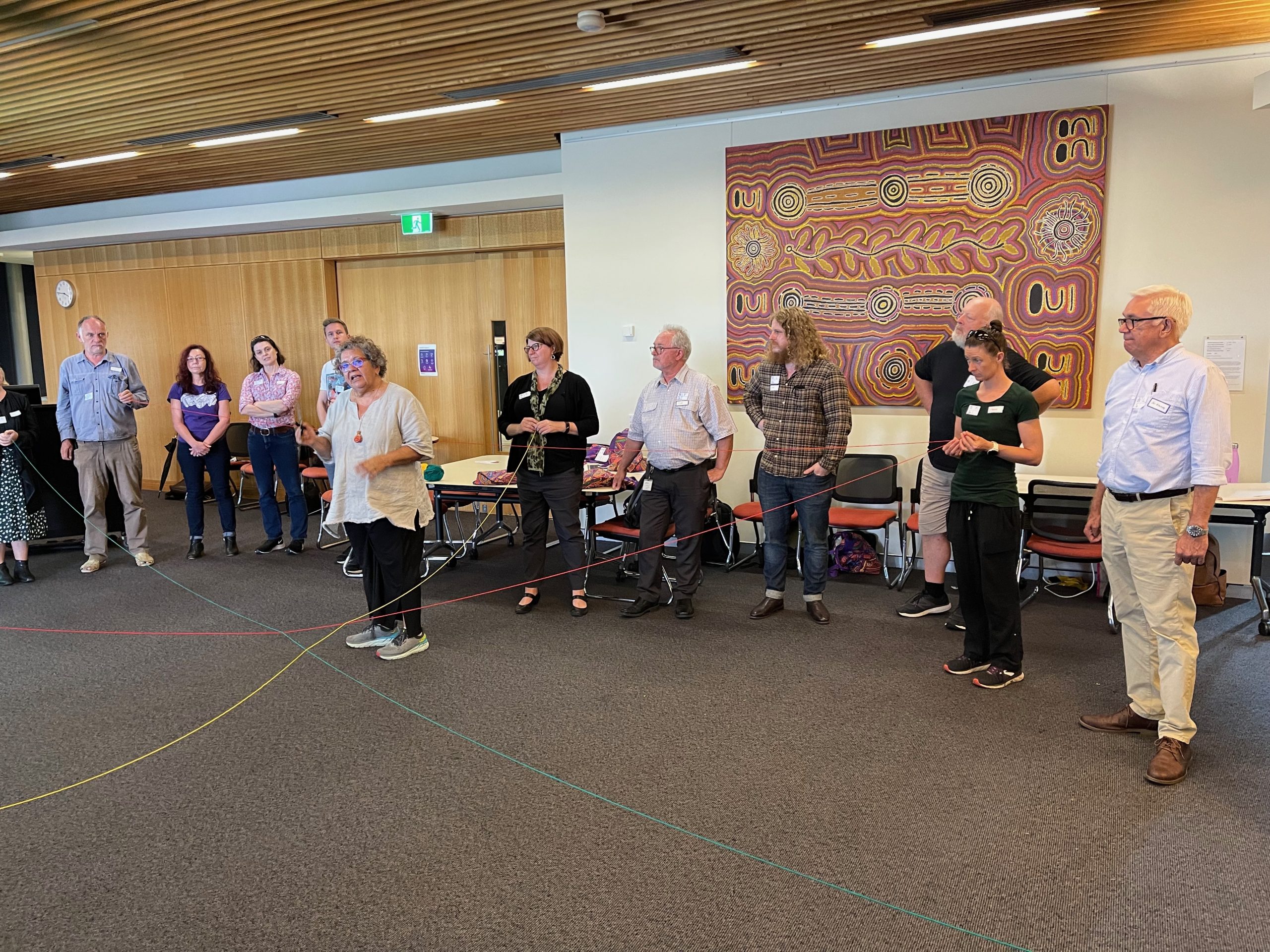 Workshop participants standing in a row behind Professor Tracey Bunda in a room. Some participants hold pieces of red, yellow and green string that cross at the centre.