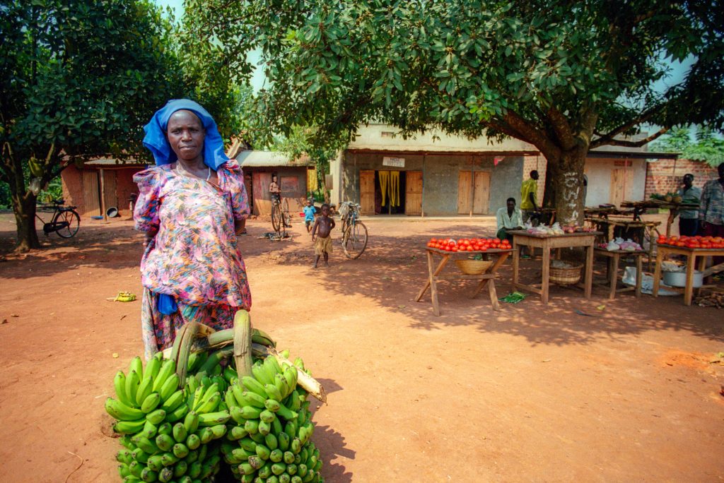 Woman with banana bunch standing in front of buildings and tables with food produce