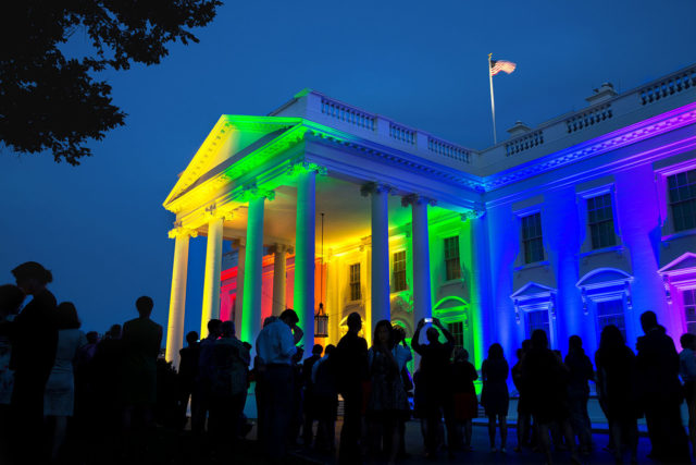 The White House at night lit up with the colours of the rainbox. People are in dark in the foreground, looking at the front of the White House, with some taking photos. The America flag waves at the top of the picture on the pole at the top of the White House.