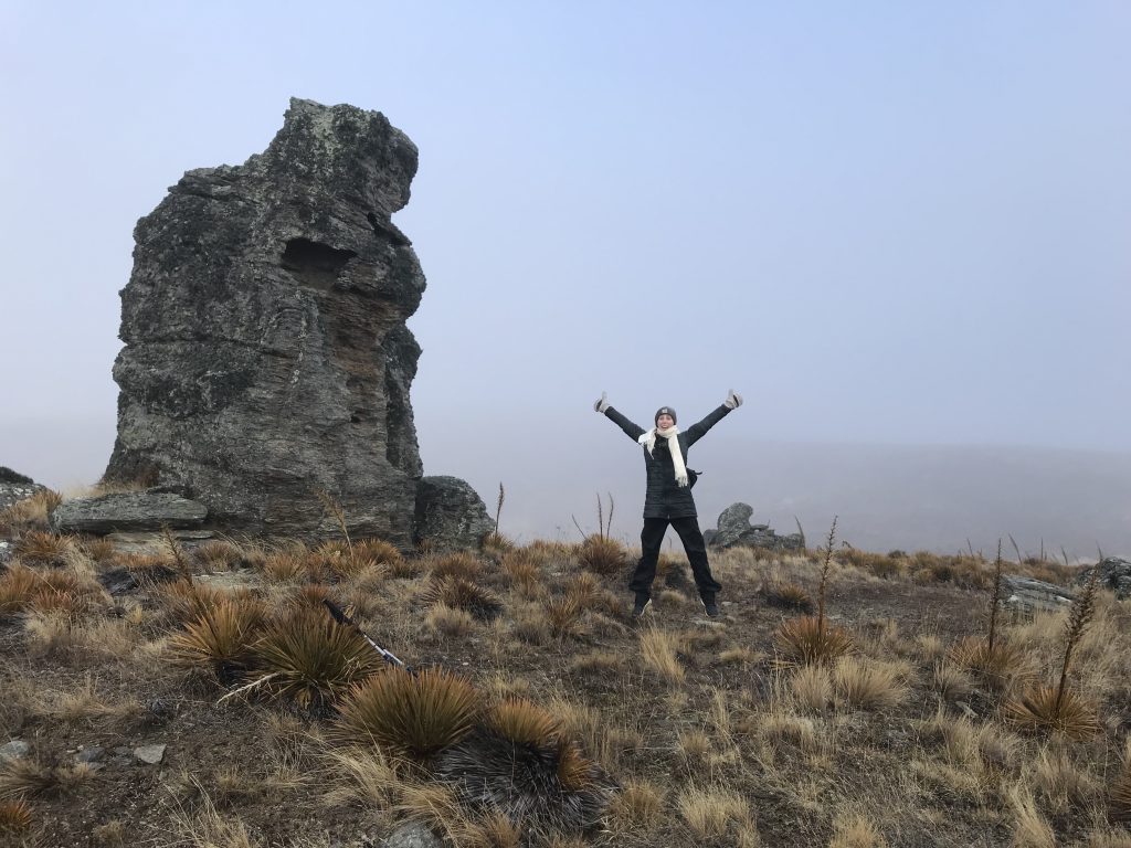 A White woman jumping in the air with her arms up while surrounded by wild grass and a large rock in the background