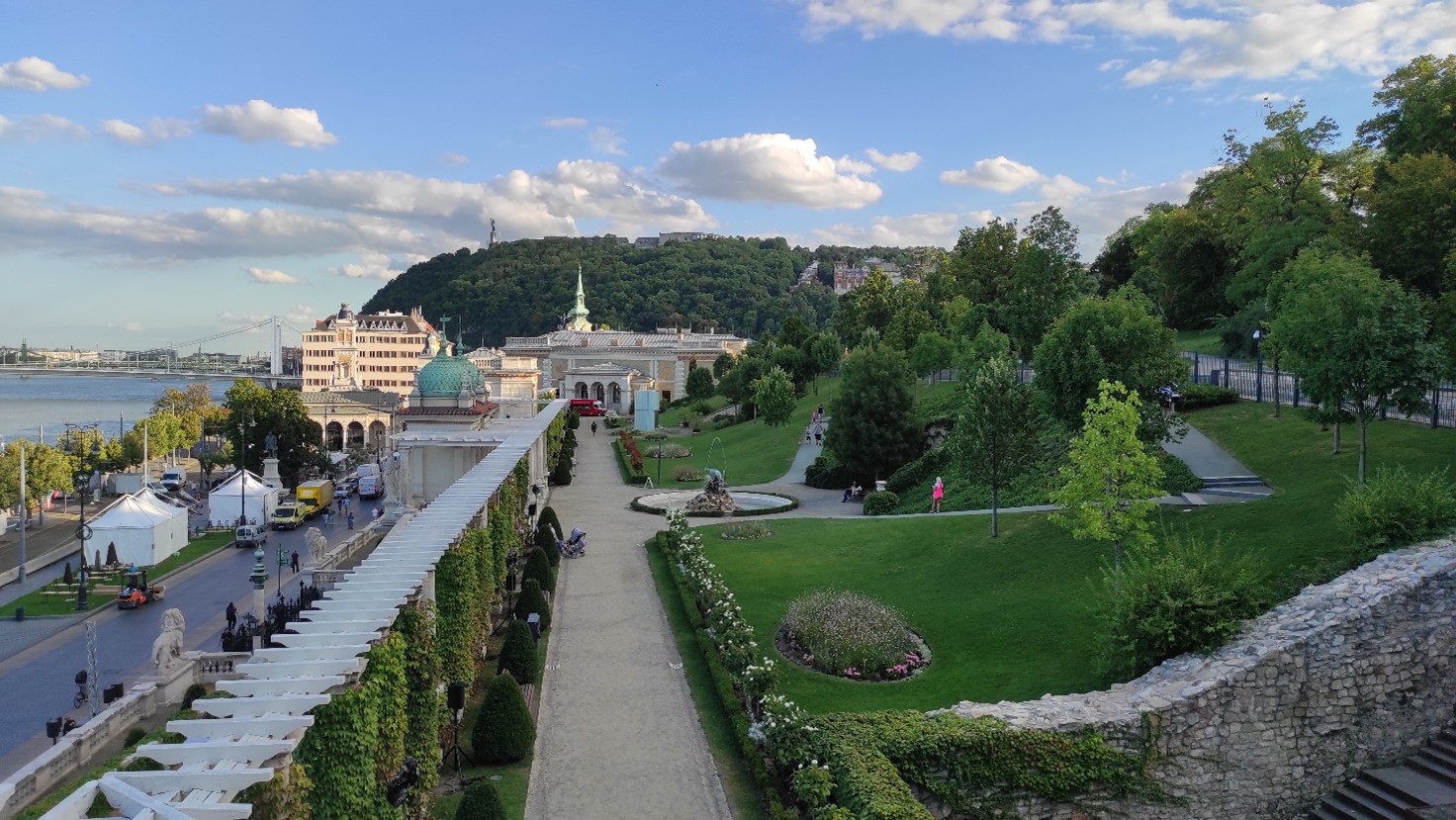 The photo is taken in the Buda side of Budapest, the camera is focussed on a hill with a statue -a female figure symbolising liberty. It’s a perspective photo with lines in the centre of the composition guiding the viewers’ attention through a park. The Danube is seen in the back. The picture has vivid colours: green, light blue and white dominate. The photo was taken during the afternoon, and it has a mackerel sky