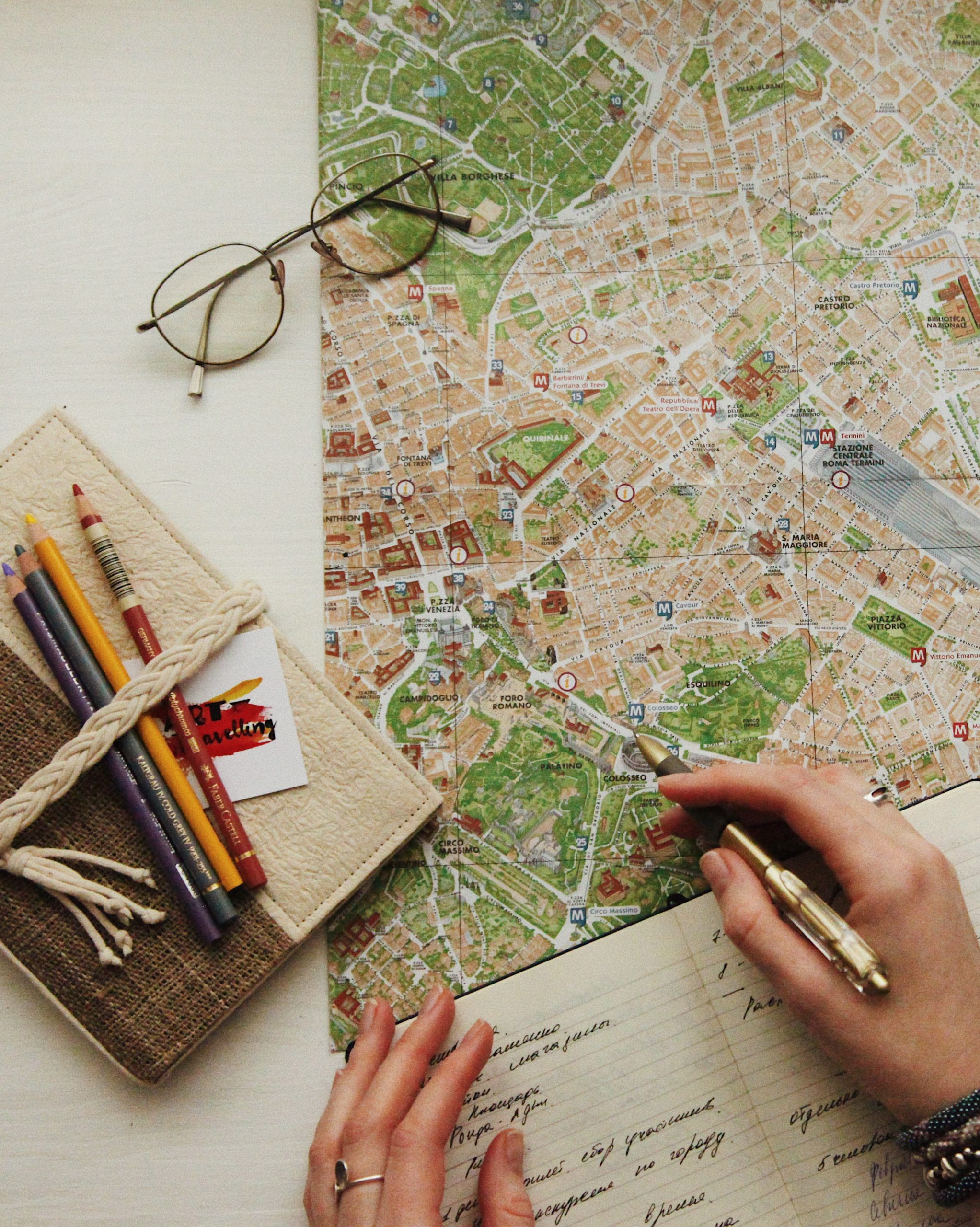 Overhead image of a person reviewing a map while writing in a open notebook. A pair of glasses, a journal, and pecils lie beside the map