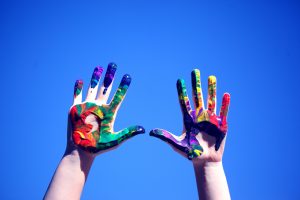 Hands covered with rainbow coloured paint held up in front of the sky.
