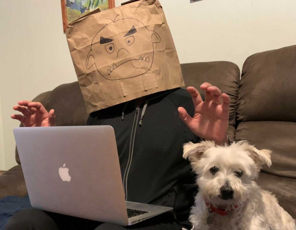 Person with head covered in paper bag with scary face on it, seated with a small dog on a couch with a laptop.